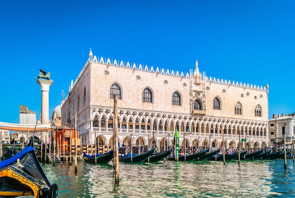 Doge's Palace in Venice, Italy, with gondolas moored in the foreground under a bright blue sky.